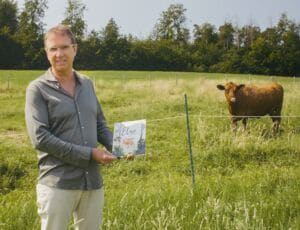 Ralph hält das Buch "Elsa büxt aus"; im Hintergrund steht Kuh Elsa