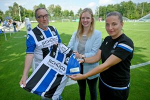 zwei Frauen und ein Mann halten ein Fußballtrikot mit CalmeMara-Ärmelpatch