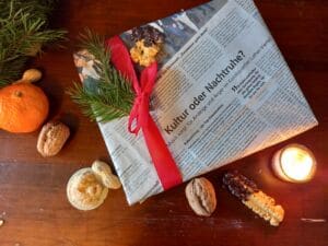ein Geschenk mit einer Zeitung als Geschenkpapier, daneben sind Nüsse, eine Orange und eine Kerze