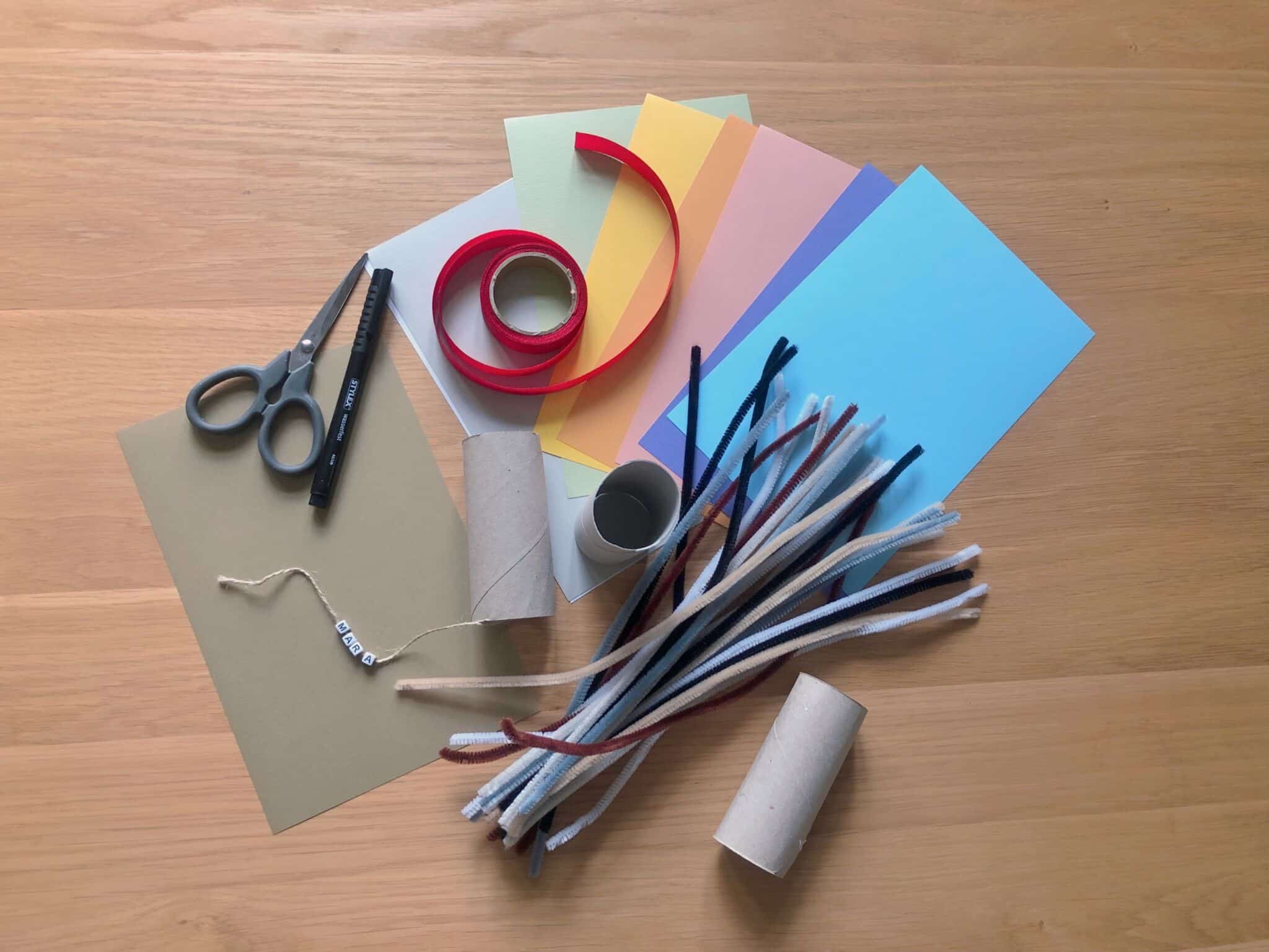 Foto: Papier, eine Schere, leere Klopapierrolle, ein Pfeifenreiniger, ein Stift und rotes Geschenkband