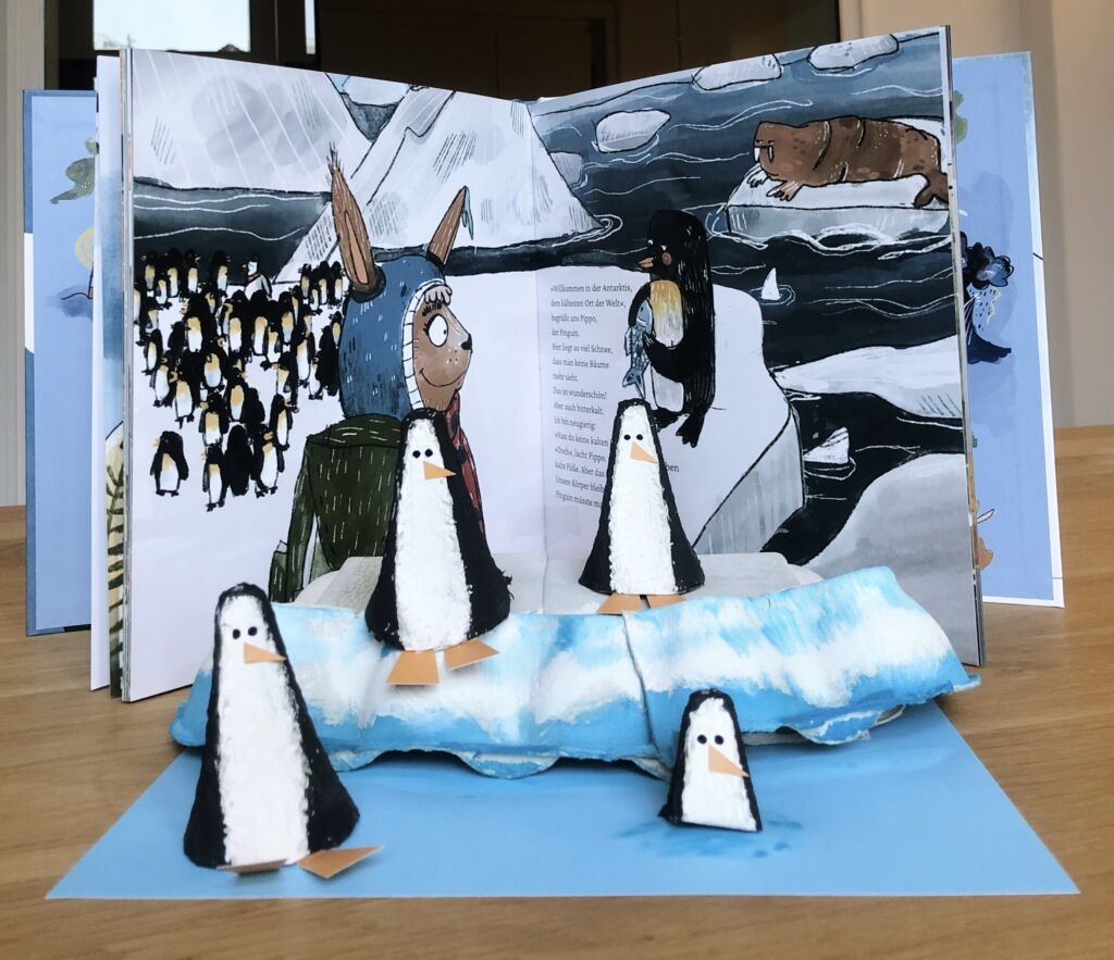 Foto: die gebastelten Penguine stehen vor dem Buch "Eine kleine große Reise"; das Buch ist aufgeklappt und zu sehen, ist eine Seite mit Pinguinen; ein paar der Pinguine stehen auf einer gebastelten Eisscholle