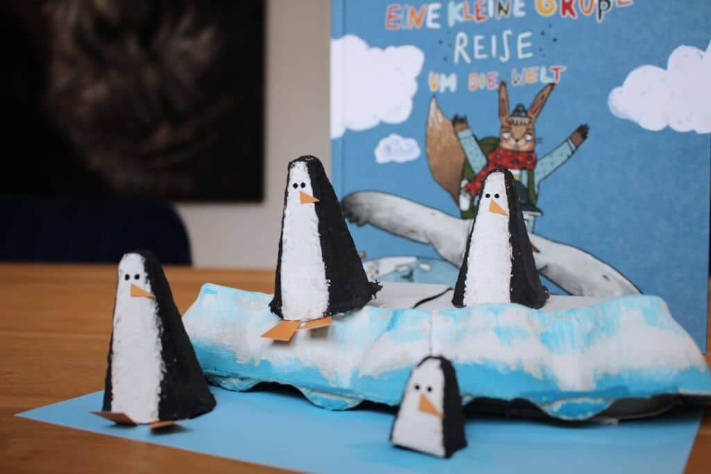 Foto: die gebastelten Penguine stehen vor dem Buch "Eine kleine große Reise", ein paar der Pinguine stehen auf einer gebastelten Eisscholle