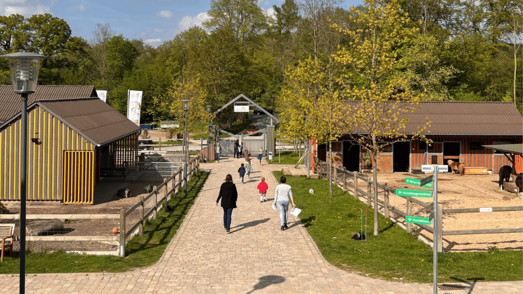 Menschen gehen auf einem Steinweg zu einem Tor mit Aufschrift "Dorf Sentana", links vom Weg ist ein Schweinestall, rechts vom Weg ist ein Pferdestall