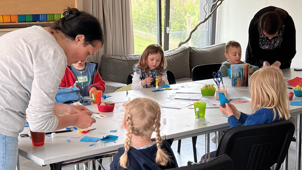 5 Kinder sitzen an einem Tisch, 2 Erwachsene stehen daneben