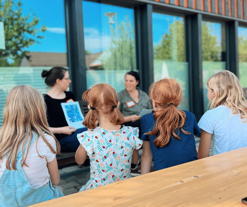 2 Frauen und 4 Mädchen sitzen; die Frauen halten Zettel oder Bücher in der Hand