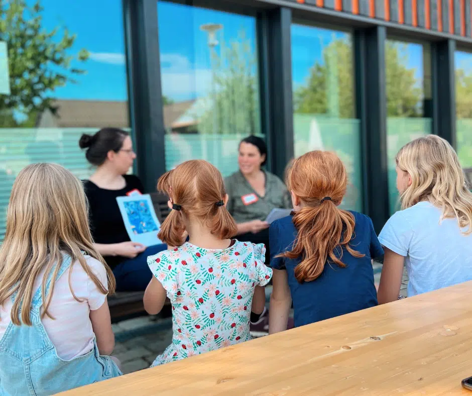 kinderlektorat2 2 Frauen und 4 Mädchen sitzen; die Frauen halten Zettel oder Bücher in der Hand
