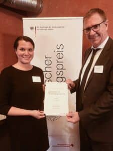 Eine Frau und ein Mann halten eine Urkunde in die Kamera. Im Hintergrund ist ein Banner zu erkennen.