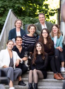 Ein Gruppe von Menschen - 1 Mann, 7 Frauen und ein kleiner Hund - sitzt auf einer Treppe. Im Hintergrund sind Bäume zu erkennnen.