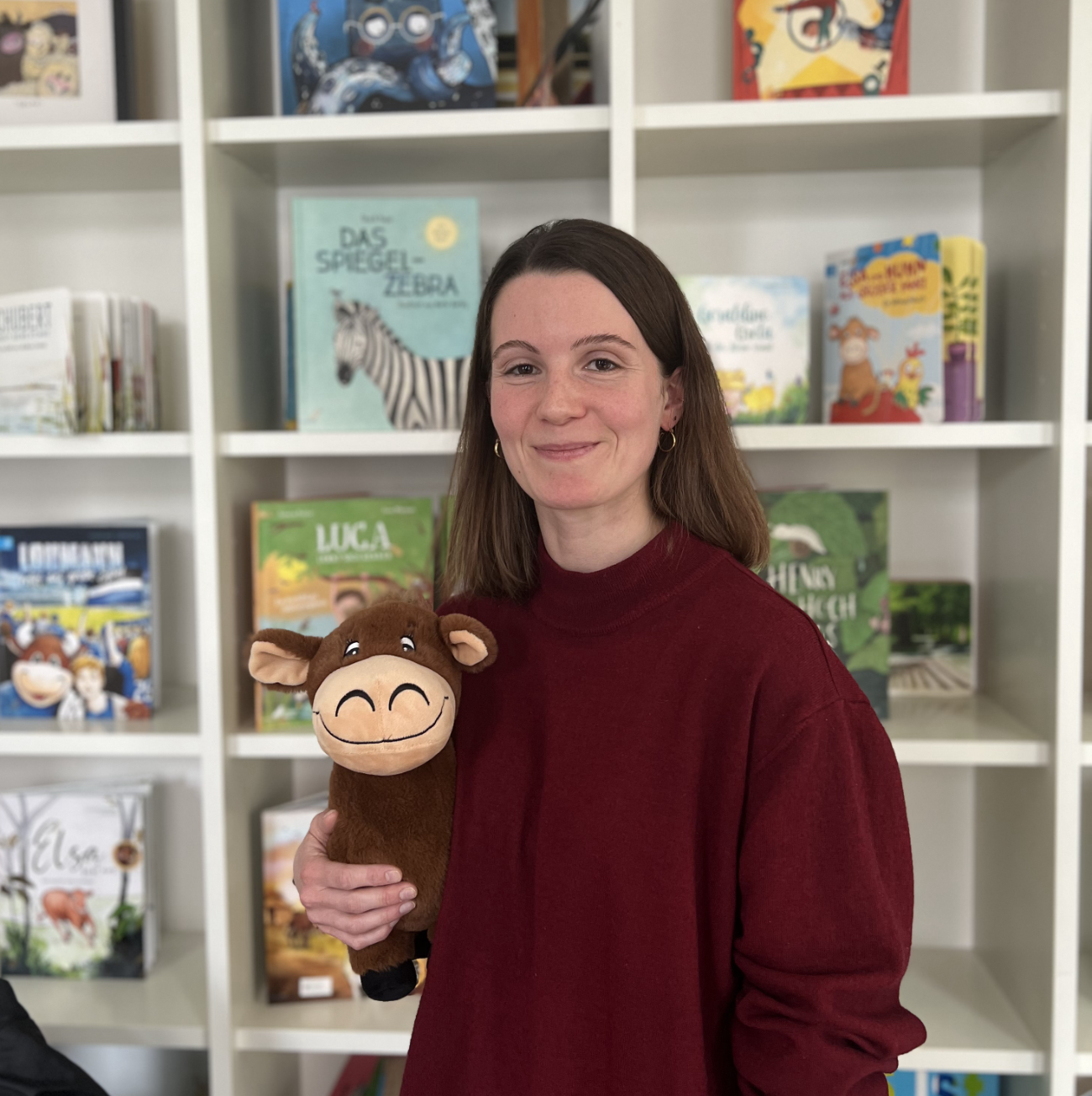 Svenja_Greifenberg_CalmeMara Svenja, eine junge Frau mit schulterlangen braunen Haaren steht lächelnd vor einem Bücherregal. Im Arm hält sie eine Kuschelkuh.