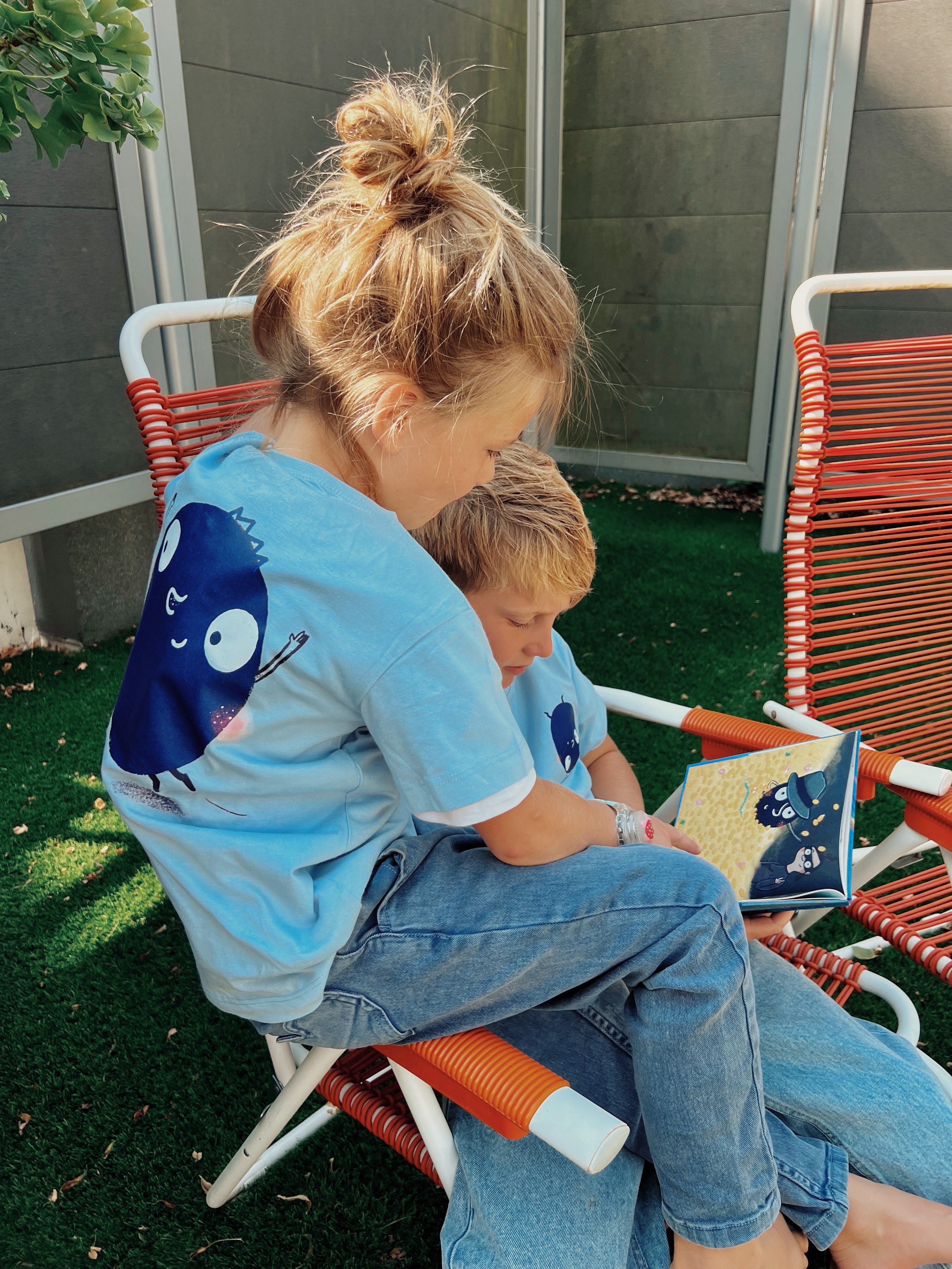 Zwei Kinder sitzen im Garten, tragen hellblaue T-Shirts mit Blaubeere-Aufdruck und lesen im Buch "Die Blaubeere"