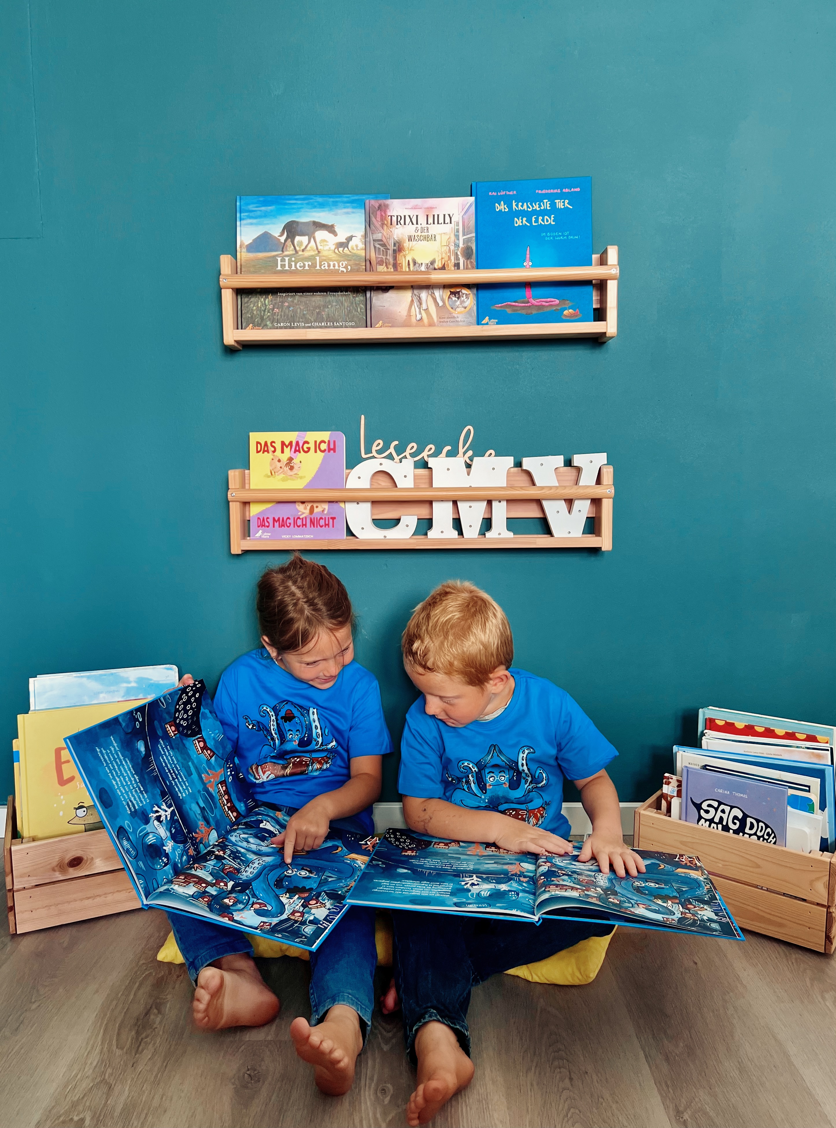 Zwei Kinder sitzen in einer Leseecke, tragen blaue T-Shirts mit Oktopus-Aufdruck und lesen im Buch "Lass es raus, Knotenklaus"