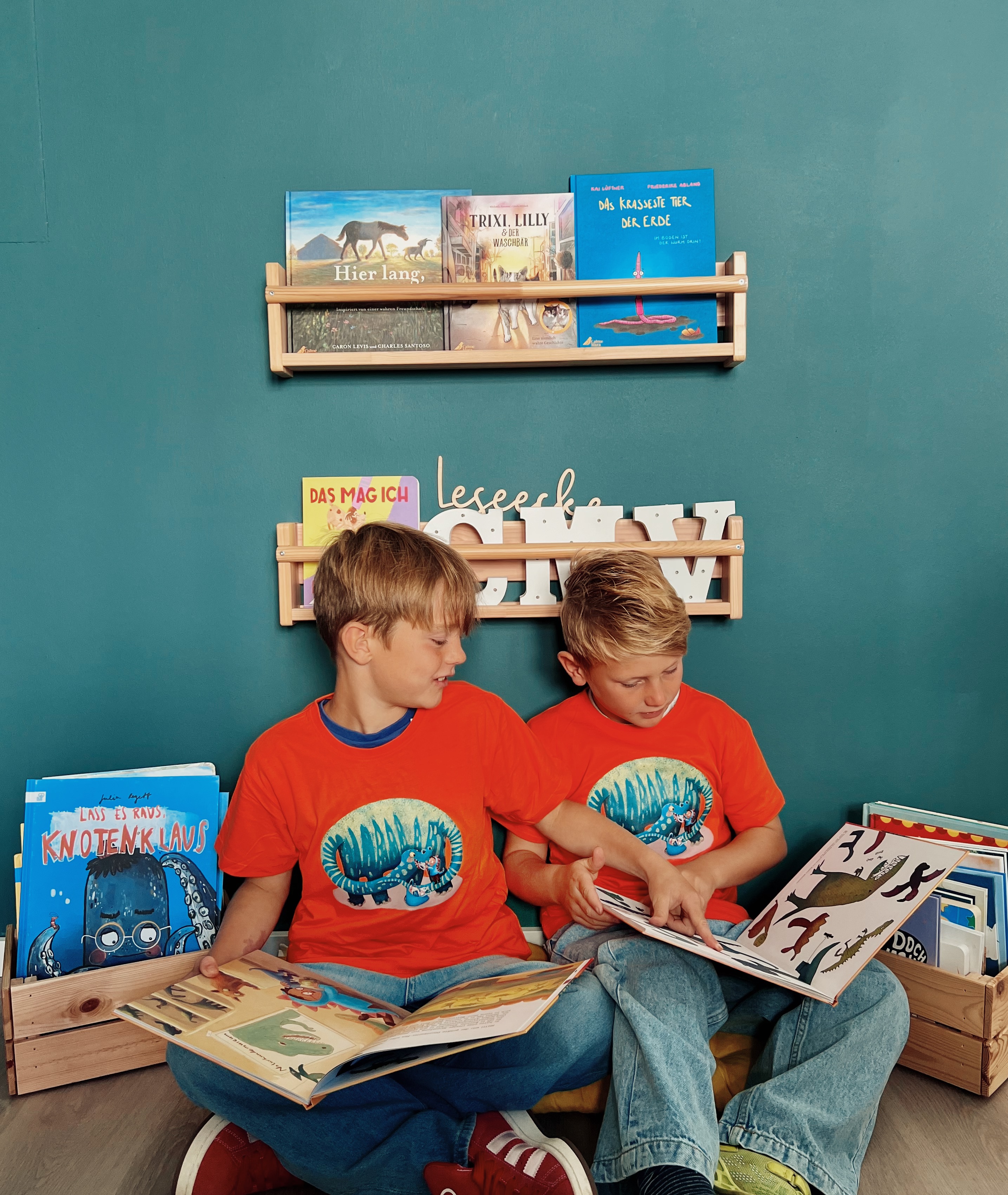 Zwei Kinder sitzen in einer Leseecke, tragen orangefarbene T-Shirts mit Dino-Aufdruck und lesen im Buch "Der Recyclosaurus"