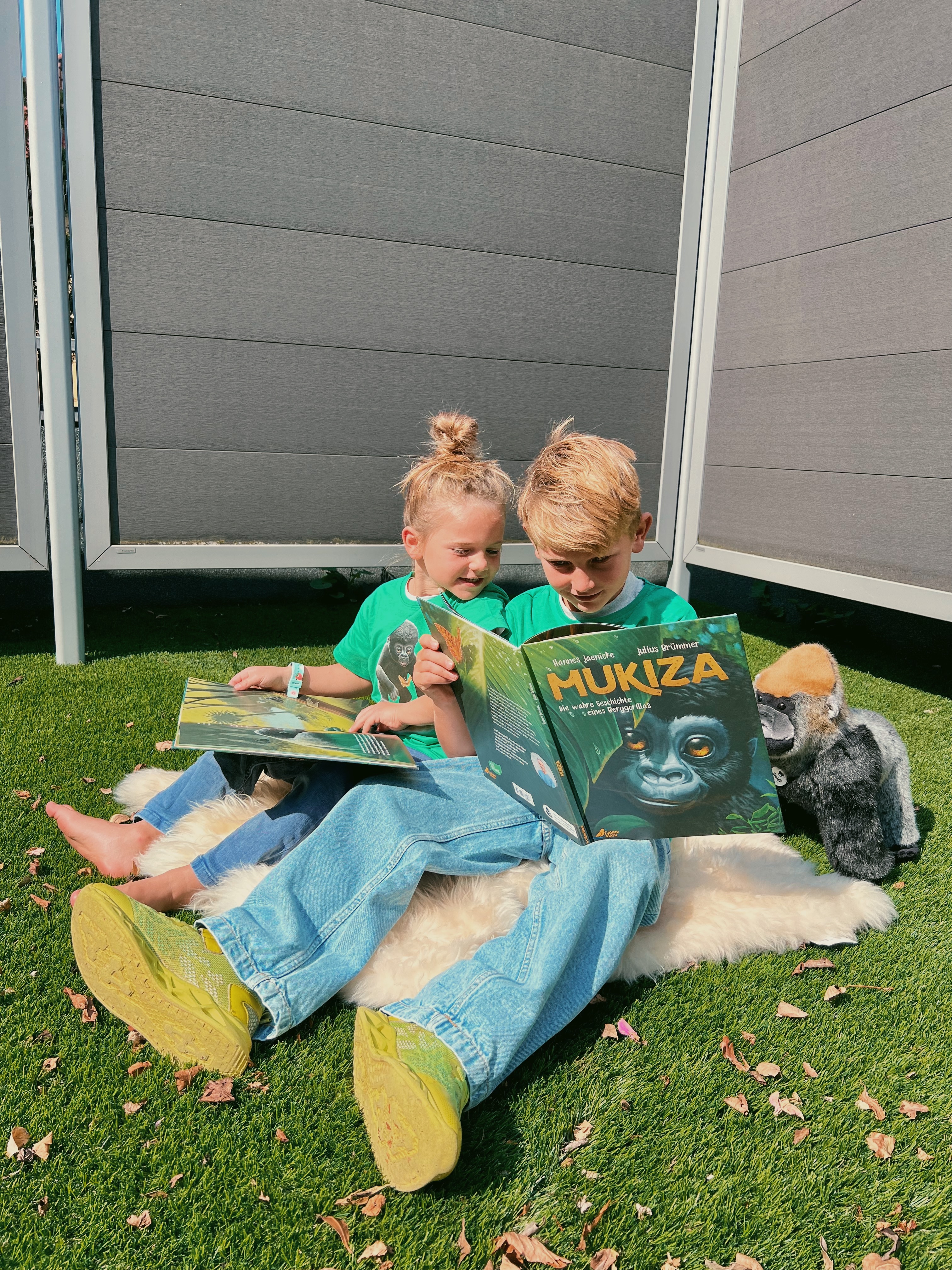 Zwei Kinder sitzen im Garten, tragen grüne T-Shirts mit Gorilla-Aufdruck, lesen im Buch "Mukiza" und spielen mit einem Stoffgorilla