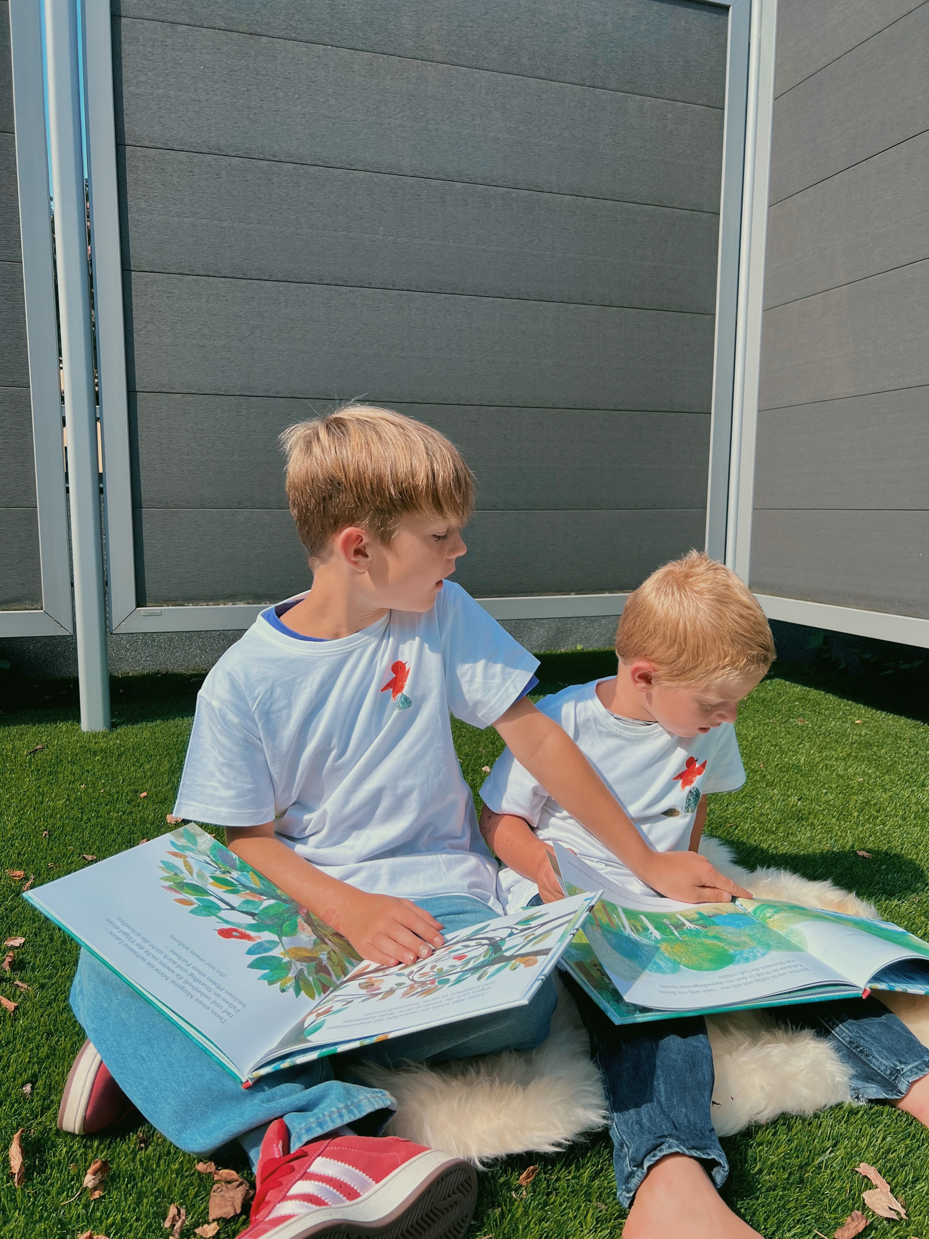 Zwei Kinder sitzen im Garten, tragen weiße T-Shirts mit Vogel-Aufdruck und lesen im Buch "Malie"