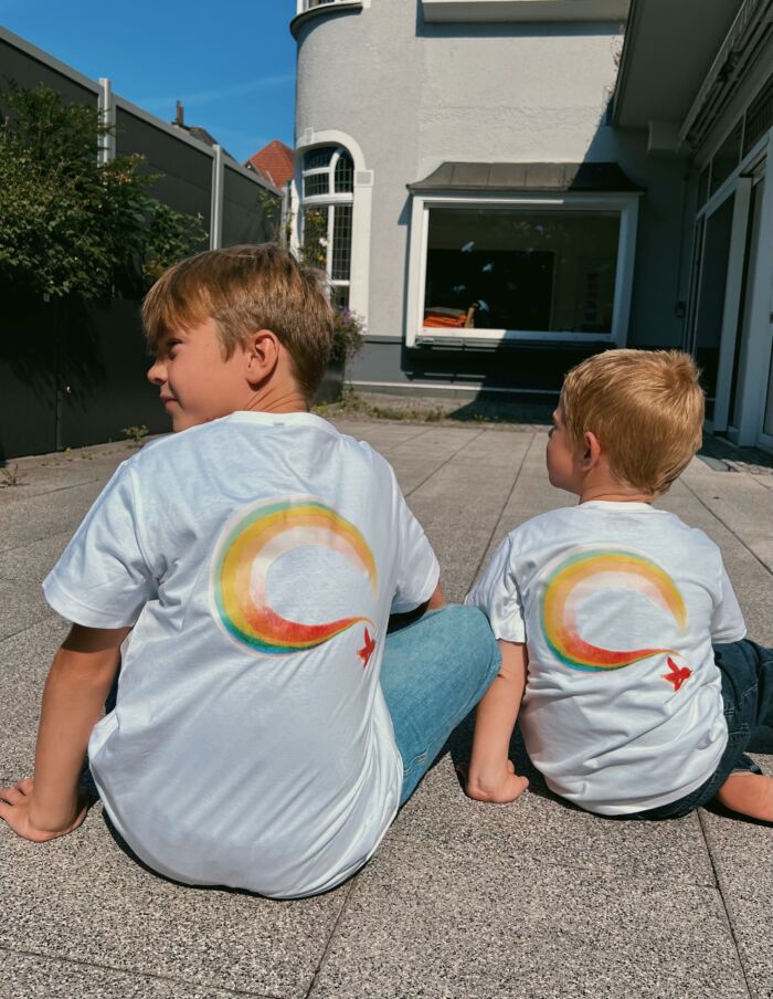 Zwei Kinder sitzen im Garten und tragen weiße T-Shirts mit Regenbogen- und Vogel-Aufdruck auf dem Rücken