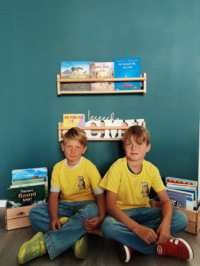 Zwei Kinder sitzen in einer Leseecke und tragen gelbe T-Shirts mit Aal-Aufdruck