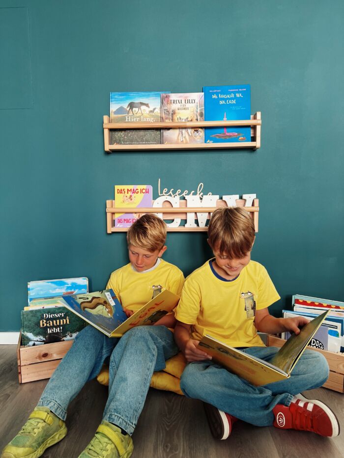 Zwei Kinder sitzen in einer Leseecke, tragen gelbe T-Shirts mit Aal-Aufdruck und lesen im Buch "Egal, sagt Aal"